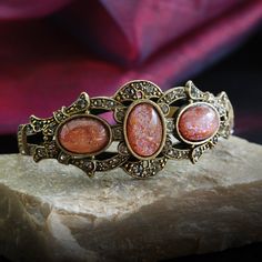 "This rich, unusually impressive bracelet is created from 90 year old stones set into our studio's original sculpture. The bracket is finely detailed and set with premium Swarovski smoke crystal. Three oval stones are vintage beauties from the 1930s which I acquired from a former exporter in Czechoslovakia. The bracket is mounted on a split shank cuff and will comfortably fit 7 to 8\" wrists. Hand burnished warm bronze finish. Entirely made in our Los Angeles studio. Choices: RUBINITE stones are Victorian Bracelet, Jelly Opal, Jewelry Cleaning Solution, Gold Flecks, Sweet Romance, Red Jewelry, Vintage Bracelet, Opal Bracelet, Old Stone