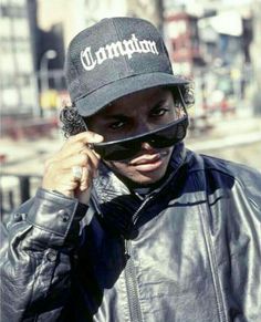 a man in a leather jacket and hat is talking on his cell phone while wearing a black baseball cap