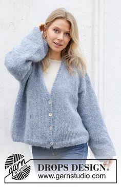 a woman wearing a blue cardigan sweater and jeans standing in front of a white wall