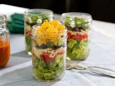 four mason jars filled with salad and dressing on top of a blue table cloth next to spoons