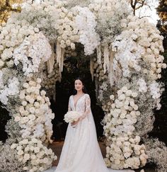 a woman in a wedding dress standing under an arch of white flowers and greenery