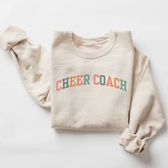 ** Please note: Orders placed after December 8 may not arrive by Christmas.  We will still do our best to fulfill all orders as quickly as possible.  We appreciate your understanding.  Happy Holidays  Introducing our cozy and stylish sweatshirt, perfect for all the amazing cheer coaches out there! Designed with a vibrant collegiate font, the words "Cheer Coach" across the chest make a bold statement that celebrates the incredible work you do. Whether you're running to practice or just relaxing a Cotton Athleisure Sweatshirt For Game Day, Varsity Sweatshirt For Cheerleading In Fall, Sports Sweatshirt With Letter Embroidery In Cotton, Sporty Cheerleading T-shirt For Fall, Cotton Sports Sweatshirt With Letter Embroidery, Sporty Long Sleeve Sweatshirt For Cheerleading, Cotton Sweatshirt With Letter Embroidery For Sports, Cotton Team Spirit Sweatshirt With Lettering, Collegiate Long Sleeve Sweatshirt For Cheerleading