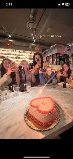 a group of women taking pictures with their cell phones while sitting at a table in front of a birthday cake