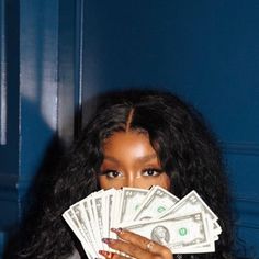 a woman covering her face with cash while standing in front of a blue wall and looking at the camera