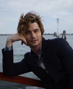 a man sitting on top of a wooden rail next to the ocean with his hand under his chin