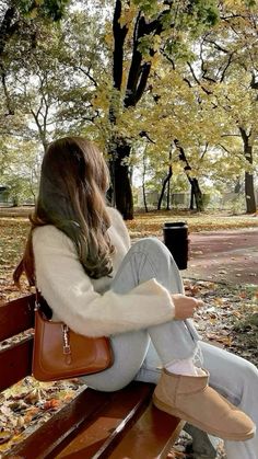 a woman sitting on top of a wooden bench in the fall leaves with her hands behind her back