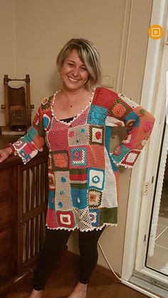 a woman standing in front of a wooden dresser wearing a multicolored crocheted sweater