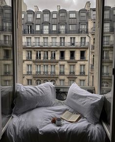 an open book and cup sit on a bed in front of a window overlooking the city