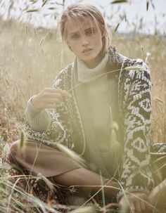 a woman sitting on the ground in tall grass wearing a sweater and pants, with her hands behind her head