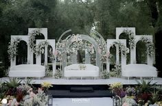 an outdoor ceremony setup with white furniture and flowers on the stage, surrounded by greenery