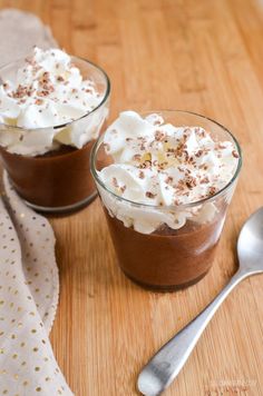 two desserts with whipped cream and chocolate on a wooden table next to a spoon