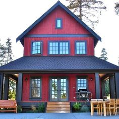 a red house with black shingles and wooden furniture outside the front door, surrounded by pine trees