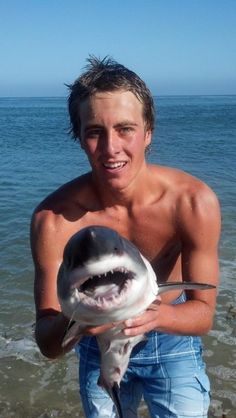 a young man holding a shark in his hands on the beach with water and blue sky behind him