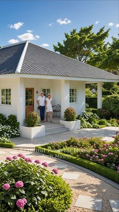 two people standing in the doorway of a white house surrounded by flowers and greenery