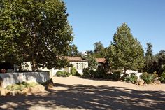 a house with trees and bushes in front of it on a dirt road next to a tree lined driveway