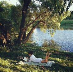 a man laying on the grass reading a book next to a lake with trees in the background