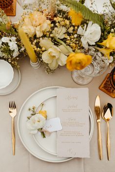 the table is set with white plates and gold cutlery, silverware, and yellow flowers