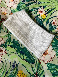 a white knitted headband laying on top of a green floral print cloth with flowers