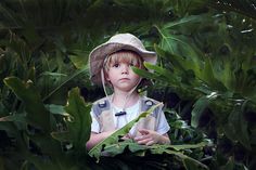 a little boy wearing a hat standing in the middle of some green plants with his hands on his chest