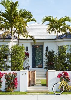 a yellow bike parked in front of a white house with red flowers and palm trees