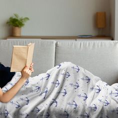 a woman laying on top of a couch under a blanket holding a book in her hand