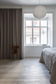 a bedroom with a bed, chair and window in the corner is lit by a hanging light