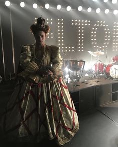 a woman dressed in an elaborate dress standing on stage with drums and lights behind her