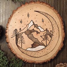 a wood slice with mountains and stars on it, sitting next to pineconi