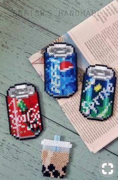 three cross stitch soda cans sitting on top of a table next to an open book