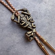 "Original scorpion design was hand carved, then cast in the Heliotrope studio in Tucson, AZ. Cast in solid brass (gold tone), or white bronze (silver tone). Cord is machine braided black or tan leather. At checkout, please choose the desired cord length: 36\"----- Recommended for people under 6' tall. 42\"----- Recommended for people 6' tall or taller. Scorpion measures 2.4\" tall x 1.5\" wide. PLEASE NOTE: final photo (AND the video) show a comparison between the LARGE SCORPION BOLO TIE (this listing), and the SMALL SCORPION BOLO TIE (see other listing)." Bohemian Hand Tooled Jewelry For Rodeo, Artisan Hand-tooled Jewelry For Rodeo, Western Engraved Jewelry For Rodeo, Western Style Engraved Jewelry For Rodeo, Hand Tooled Brown Jewelry For Rodeo, Brown Hand Tooled Jewelry For Rodeo, Vintage Gold Jewelry For Rodeo, Handmade Bohemian Bolo Tie For Rodeo, Handmade Western Jewelry For Rodeo