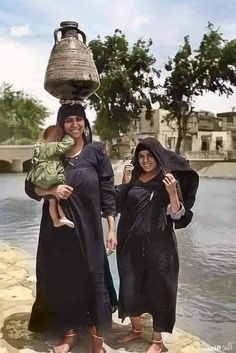 two women in black dresses holding a baby and an oil pot on top of their head