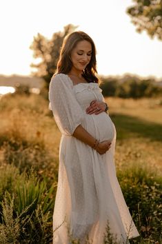 a pregnant woman in a white dress standing in tall grass with her hands on her belly