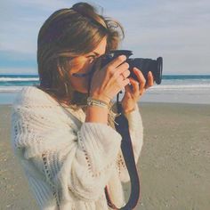 a woman is taking a photo on the beach with her camera and wearing a white sweater