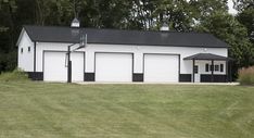 a white barn with black roof and two garages on the grass in front of trees