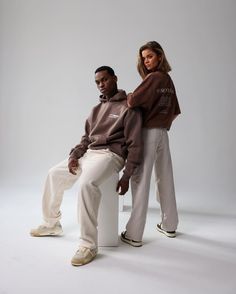 a man and woman sitting on a white stool in front of a gray background, both wearing brown sweatshirts