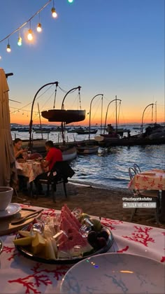 a table with food on it and people sitting at tables near the water in front of boats