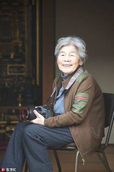 an old woman sitting on a chair with a camera in her hand and smiling at the camera