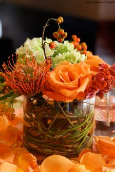 an arrangement of flowers in a glass vase on a table with orange napkins and glasses