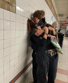 two people hugging each other while standing next to a wall with tiled walls in the background