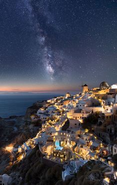 the night sky is filled with stars and buildings on top of a hill overlooking the ocean