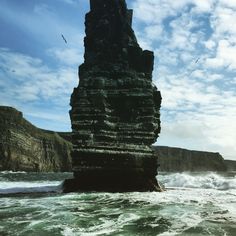 a large rock sticking out of the ocean next to a rocky cliff face with waves crashing on it