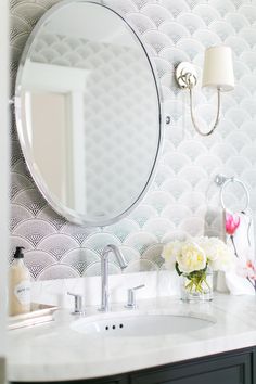 a bathroom sink with a round mirror above it and flowers in a vase on the counter