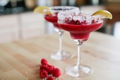 two glasses filled with raspberry margaritas on top of a wooden table and garnished with lemon wedges