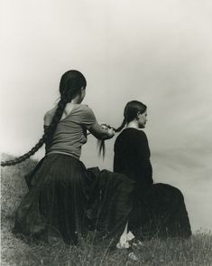 two women sitting on top of a grass covered hill with long hair in ponytails