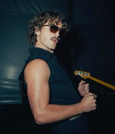 a young man with curly hair and sunglasses holding a guitar in his right hand while looking at the camera