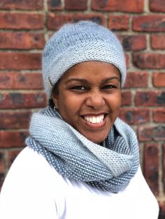 a smiling woman wearing a knitted hat and scarf in front of a brick wall