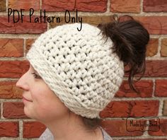 a woman wearing a white crochet beanie against a brick wall with her hair in a ponytail