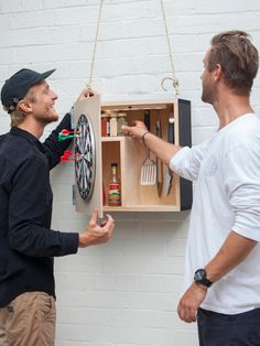 two men standing next to each other in front of a wall mounted dart and darts