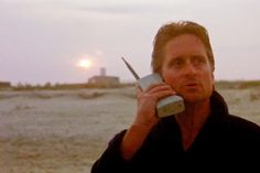 a man holding a cell phone up to his ear while standing on the beach at sunset