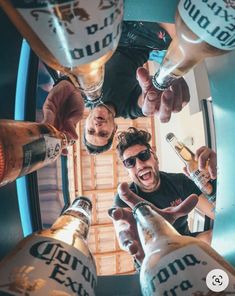 four men are standing in a circle holding beer bottles and pointing at the camera with their hands